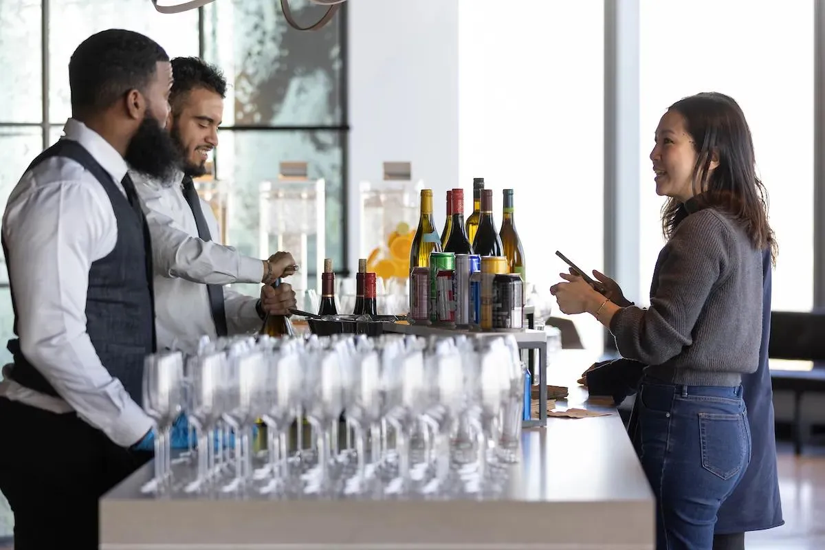 A woman standing at a bar filled with champagne glasses and wine bottles at an event.