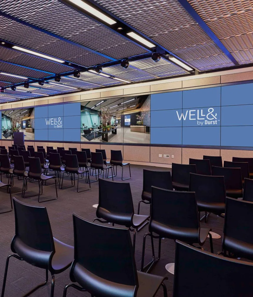 151 West 42nd conference space filled with empty chairs and a large screen that extends the far end of a wall. It displays the "Well& by Durst" logo on it.