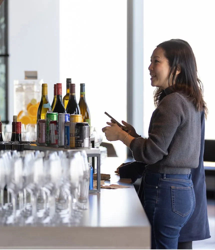 A woman standing at a bar filled with champagne glasses and wine bottles at an event.