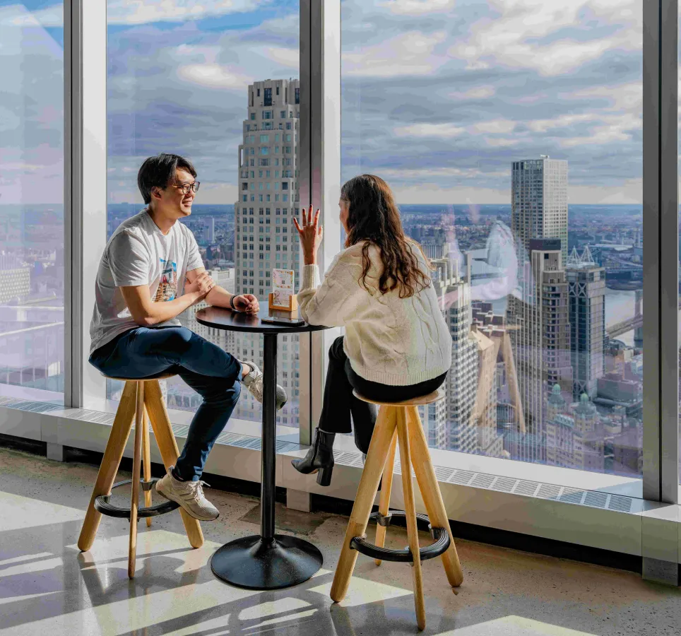A man and woman sitting at a coffee table against a window paned wall. Outside the window is New York's iconic cityscape.