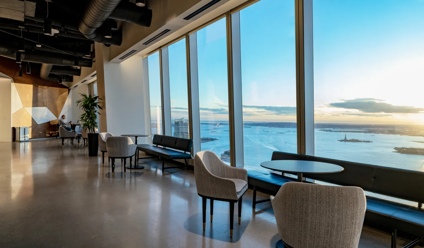 64th Floor of One World Trade Center. Modern office lounge with large windows offering a panoramic view of a city's waterfront at sunset, furnished with sofas and armchairs.