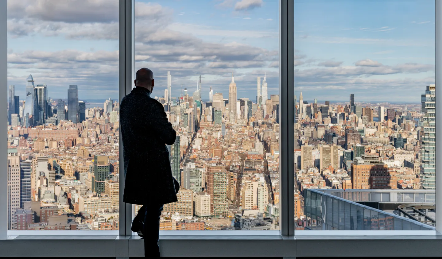 A man gazing out a window at a city skyline from the 64th floor sky lobby of One World Trade Center.