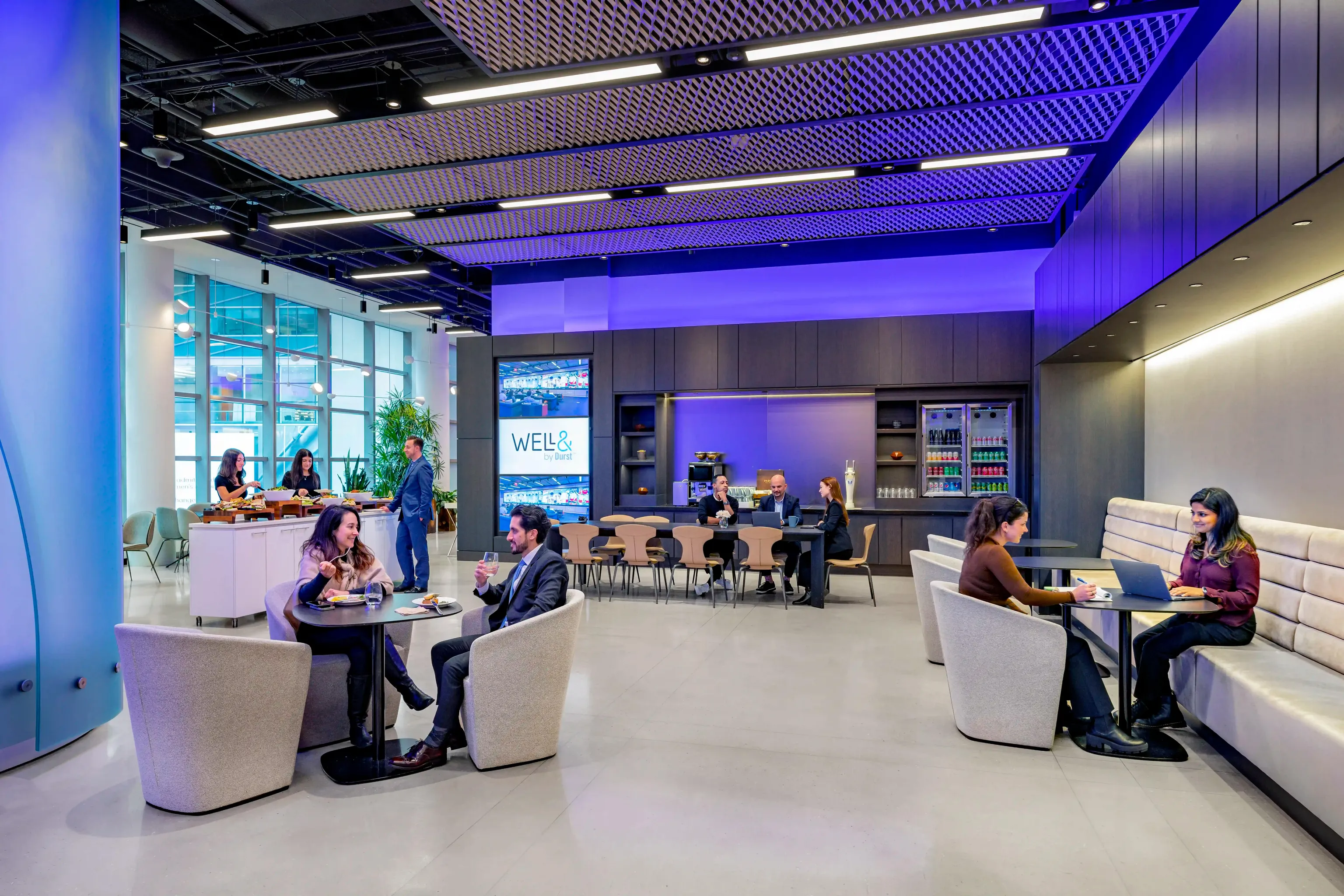 An auditorium or conference space filled with empty chairs and a large screen that extends the far end of a wall. It displays the "Well& by Durst" logo on it.