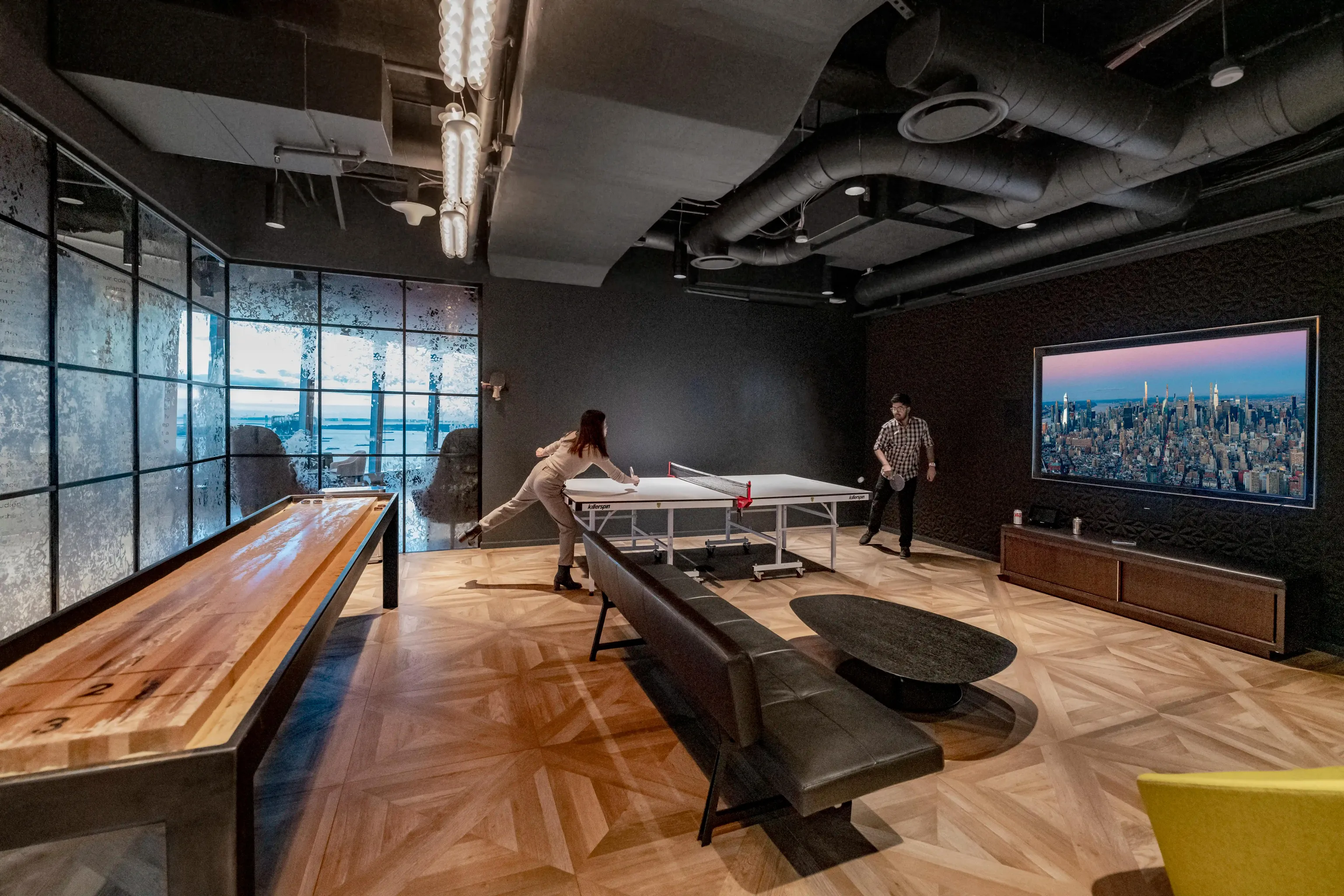 Two people in a grey walled game room are playing ping pong. The room has wooden floors, a dark grey couch and a wide screen tv hanging on the wall.