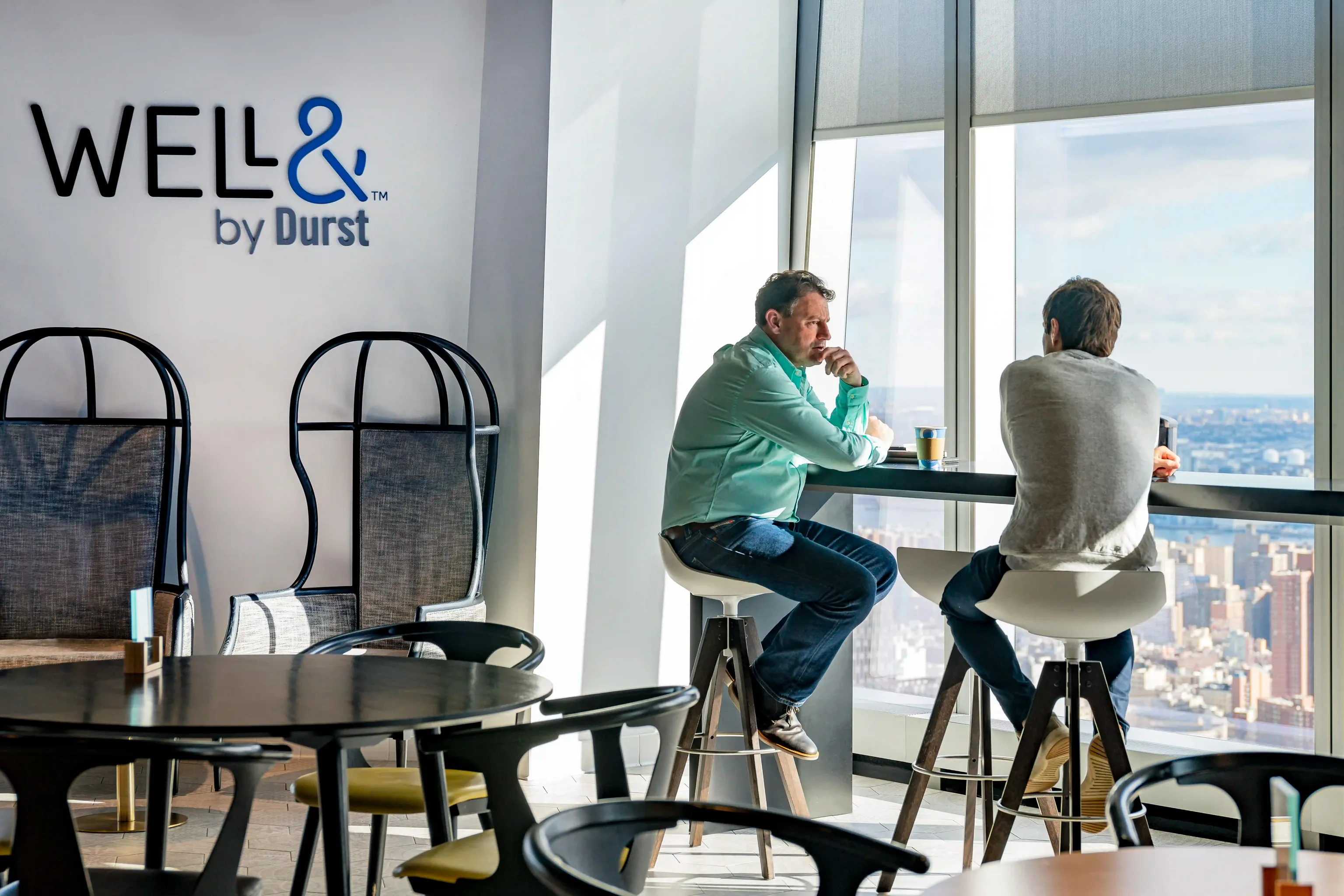 Two men sit on stools by a large window overlooking the city with a view of New York City. There is a Well& by Durst logo logo on the wall next to them.