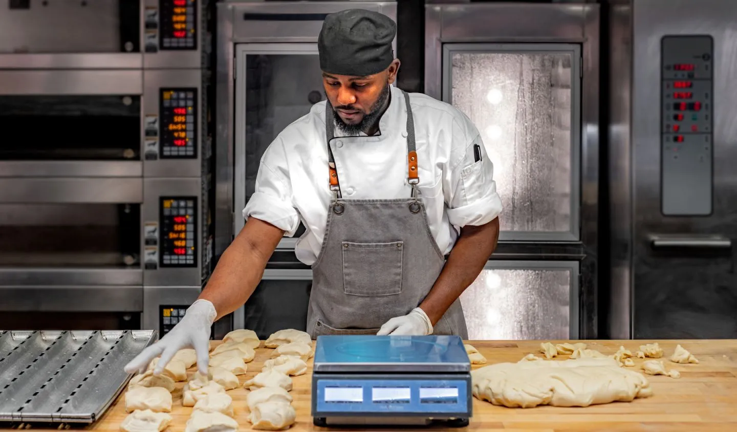 A man in an apron weighing dough portions with ovens in the background.